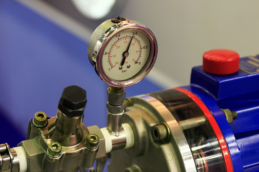Manometr wskazujący ciśnienie w instalacji hydraulicznej, zamontowany na metalowej armaturze, z widocznym elementem pompy w tle.