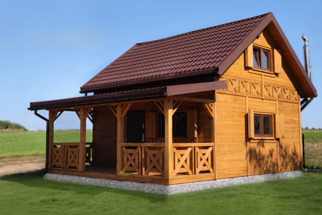 Drewniany domek letniskowy z poddaszem i werandą, z czerwonym dachem, stojący na zielonej trawie, w otoczeniu otwartej przestrzeni na tle czystego, niebieskiego nieba.