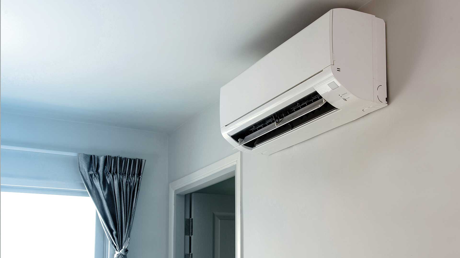 Klimatyzator typu split zamontowany na ścianie w jasnym pomieszczeniu, nad drzwiami i obok okna z zaciągniętymi zasłonami.