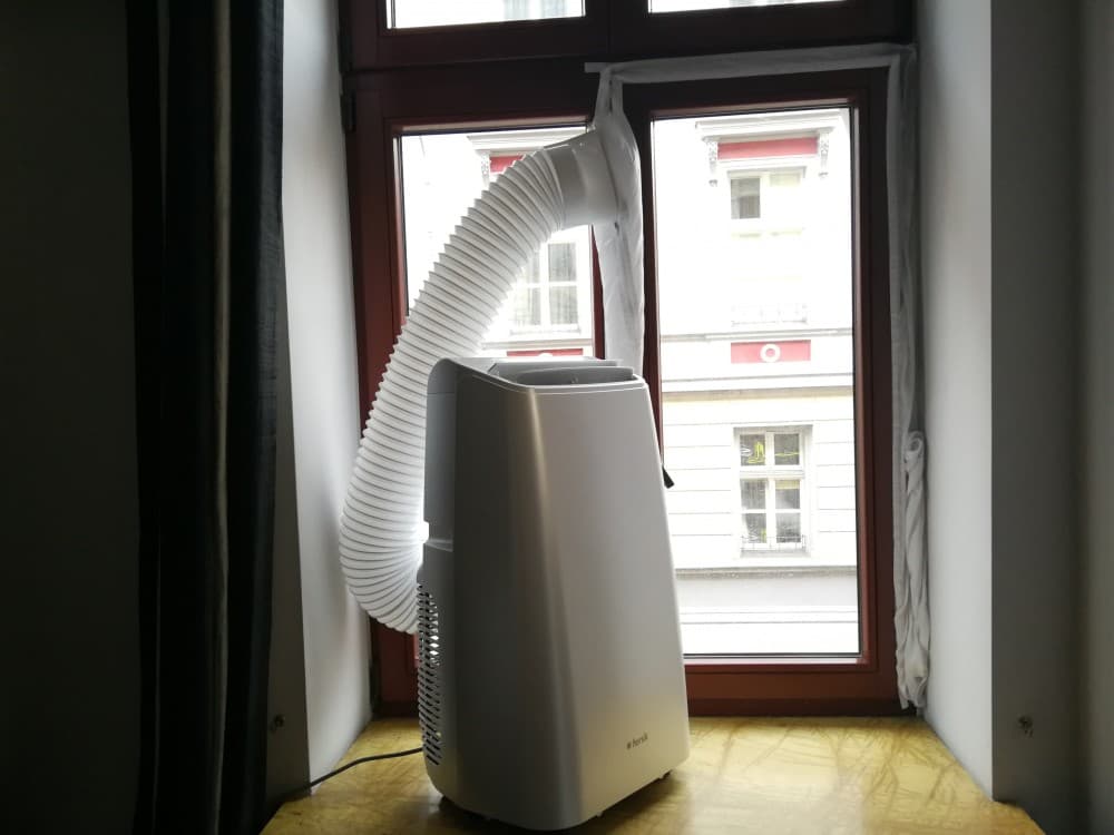 Klimatyzator przenośny, umieszczony pod oknem z wyprowadzoną rurą wentylacyjną przez okno.