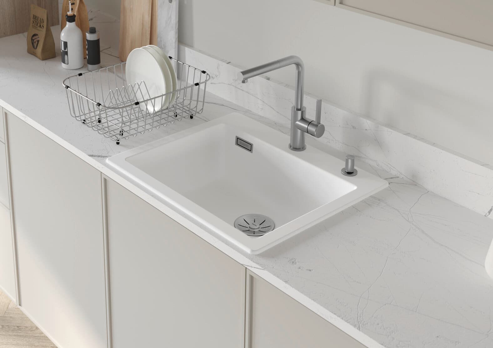 Nowoczesny, biały zlewozmywak ceramiczny wkomponowany w marmurowy blat kuchenny. Obok zlewu znajduje się stalowy ociekacz z naczyniami, a nad zlewem elegancka, metalowa bateria. W tle stoją kuchenne akcesoria, takie jak deski do krojenia oraz butelki z pr